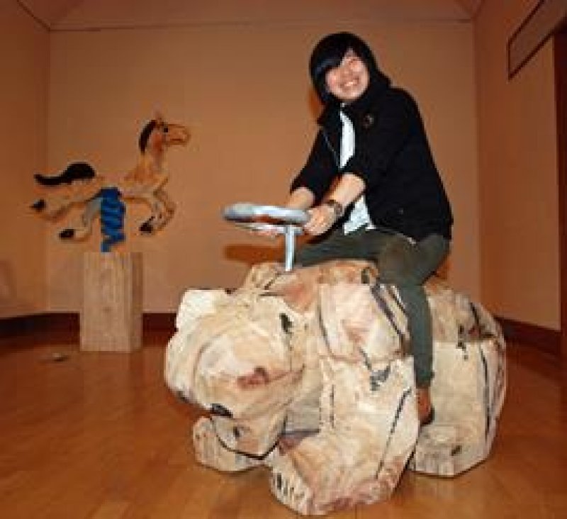 ふなばしアンデルセン公園・子ども美術館で開かれている企画展「ものごころ展」で、作品「ものモノ　屋上の乗れる熊」に座る木彫家の山本さん。後方の作品は「ものモノ　公園の乗れない馬」