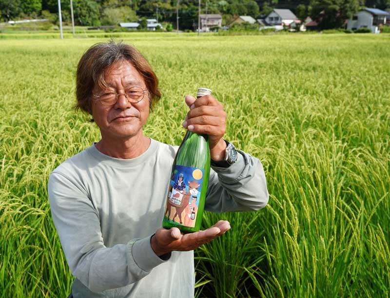山田錦の田んぼの前で日本酒「チャレンジＯＮＪＵＫＵ」を手にする吉野代表理事＝御宿町
