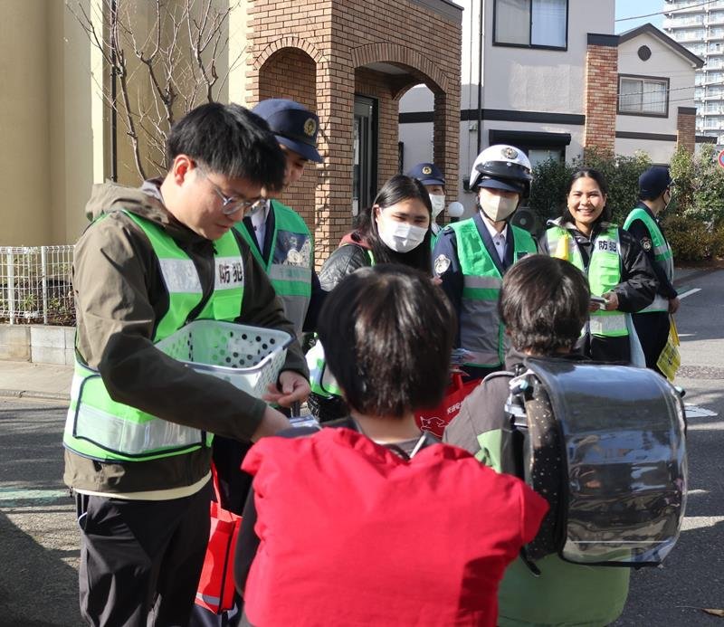交通安全グッズを児童に配り、下校を見守った外国人留学生と千葉中央署員＝２３日昼、千葉市中央区の院内小学校前
